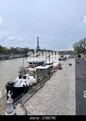 Fluss seine mit Menschen, die auf dem gepflasterten Weg spazieren. Flussboote im Vordergrund und der Eiffelturm und der Pont Invalides im Hintergrund. Stockfoto