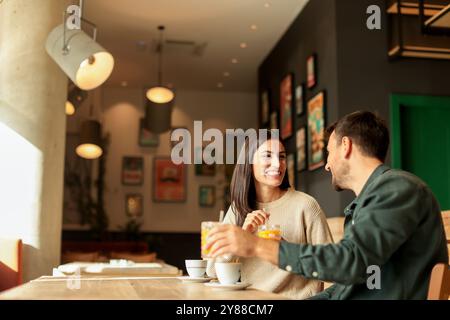 Ein Paar teilt sich einen freudigen Moment in einem stilvollen Café, schlürft Kaffee und genießt hellen Orangensaft, während es gemeinsam lacht, umgeben von einem einladenden Restaurant Stockfoto