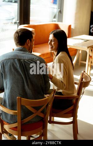 Ein Paar teilt sich einen freudigen Moment in einem Café und schlürft erfrischende Getränke, während er an einem Holztisch sitzt, umgeben von warmem Sonnenlicht und einem einladenden atmos Stockfoto