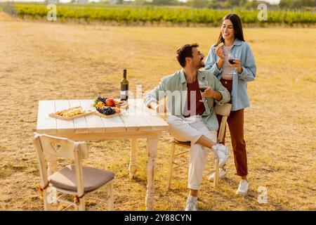 Ein paar entspannen sich an einem rustikalen Tisch in einem sonnigen Weinberg, genießen Wein und Gourmet-Snacks. Sie teilen Lachen und Freude, umgeben von üppigen Weinstöcken und dem Stockfoto