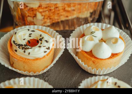 Die Torten mit Mango und Torten mit Dorkkäse sind in einer Reihe auf schwarzem Teller angeordnet. Vitrine mit einer Vielzahl von Kuchen, Cupcakes und Desserts. Stockfoto