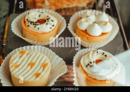 Vitrine mit einer Vielzahl von Kuchen, Cupcakes und Desserts. Die Torten mit Mango und Torten mit Dorkkäse sind in einer Reihe auf schwarzem Teller angeordnet. Stockfoto
