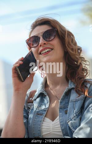 Porträt einer jungen Frau mit Sonnenbrille, die draußen auf dem Handy spricht Stockfoto