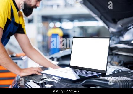 Modell-Laptop im Auto mit geöffneter Motorhaube, während Mechaniker im unscharfen Hintergrund den Kraftstofftank ersetzt. Isoliertes Bildschirmgerät neben akribischem Garagenarbeiter, der das Kundenfahrzeug repariert Stockfoto