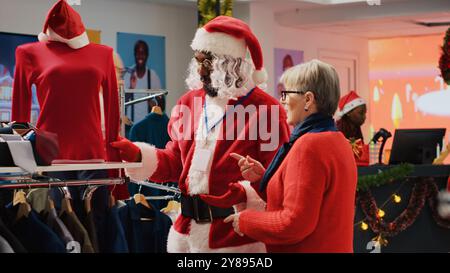 Angestellte mit Weihnachtsmann-Kostüm und -Brille hilft älteren Kunden im Geschäft, rote Bluse für Weihnachtsfeiern zu finden. Die Arbeiterin hilft älteren Frauen während der Weihnachtsferien im Modegeschäft Stockfoto