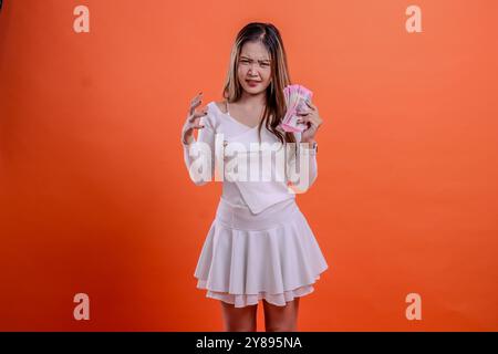 Porträt der schönen erwachsenen Frau in der weißen Spitze stehend genervt irritierte Hand, die Rupiah Bargeld hält und Hand packend Mund isolierter oranger Hintergrund. Stockfoto