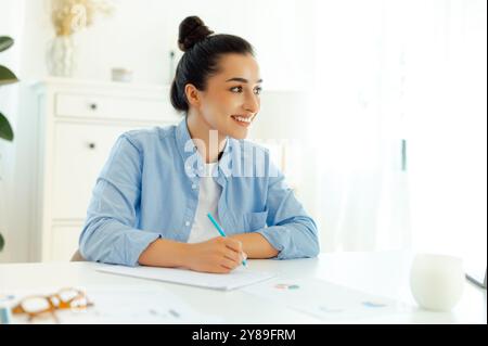 Positive charmante weiße brünette junge Frau in einem blauen Hemd, sitzt am Schreibtisch, Mitarbeiter des Unternehmens, schreibt Informationen, bereitet Präsentation vor, schaut weg, denken, lächeln Stockfoto