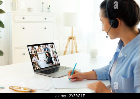 Online-Vortrag im Finanzbereich. Seitenansicht einer kaukasischen Frau sitzt an einem Schreibtisch, wo sie sich online mit multirassischen Menschen und Tutoren trifft, Unterricht hört, ihr Wissen verbessert, Notizen macht und ein Brainstorming in der Gruppe führt Stockfoto