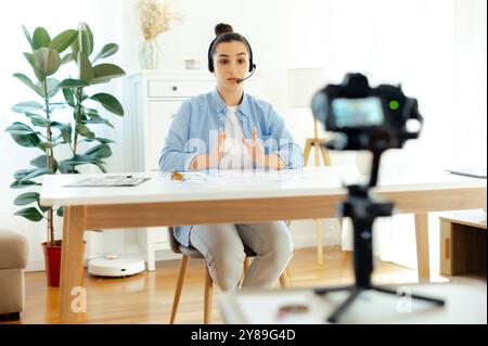 Online-Webinar, Podcast aufzeichnen. Hübsche weiße brünette junge Frau, Coach, Blogger, Influencer sitzt an einem Schreibtisch, schaut in die Kamera, nimmt Ausbildung Tutorial Vlog, Video Kurs für soziale Medien Stockfoto