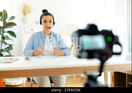 Selbstbewusste hübsche indische oder arabische brünette junge Frau, Blogger, Influencer sitzt an einem Schreibtisch, schaut sich die Kamera an, nimmt Lernvlog auf, Videokurs für soziale Medien Stockfoto