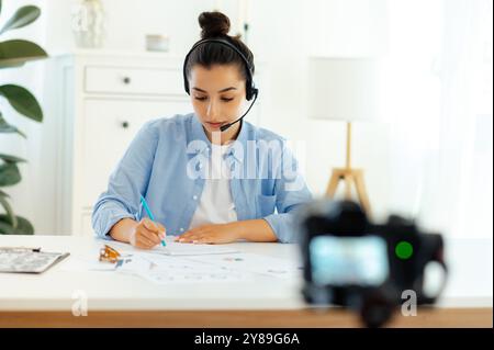Fokussierte hübsche kaukasische brünette junge Frau, Blogger, Influencer sitzt an einem Schreibtisch, Aufzeichnung von Bildung Tutorial Vlog, Video Kurs für soziale Medien, Schreiben von Informationen Stockfoto