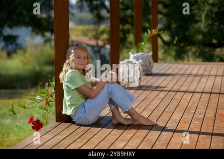 Ein junges Mädchen mit einem Lächeln in Jeans und grünem Oberteil, das auf einer Terrasse sitzt Stockfoto