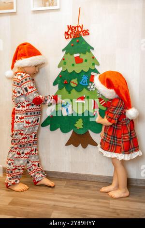 Zwei Kinder mit Weihnachtsmützen schmücken einen Weihnachtsbaum mit farbenfrohen Ornamenten. Die Umgebung ist fröhlich und festlich, sehr hell Stockfoto