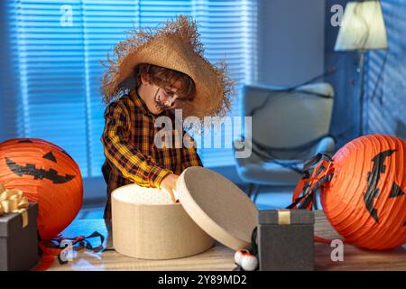 Süßer Junge, der wie Vogelscheuche gekleidet ist, mit festlichem Dekor und Geschenkboxen drinnen in der Nacht. Halloween-Feier Stockfoto