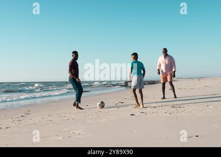 afroamerikanischer Junge spielt Fußball mit Vater und Großvater am Strand vor klarem Himmel Stockfoto