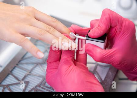 Eine Manikürerin in rosa Handschuhen trägt sorgfältig die Basis für die gelpolierung auf die Nägel eines Kunden in einem Salon auf. Stockfoto