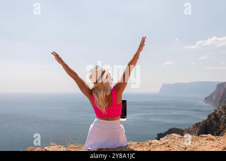 Eine Frau sitzt auf einem Hügel mit Blick auf das Meer mit einem Laptop im Schoß. Sie trägt ein rosafarbenes Oberteil und einen Strohhut. Konzept der Entspannung und Stockfoto