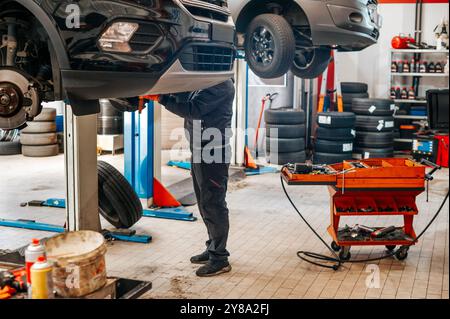 Der Mechaniker arbeitet unter einem Fahrzeug, das auf einer hydraulischen Hebebühne angehoben wird, und repariert den Motor in einer Werkstatt Stockfoto