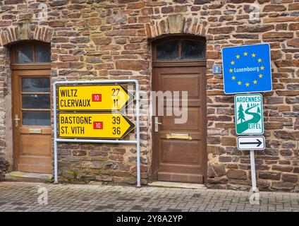 Gelbe Wegweiser nach Wiltz, Clervaux, Bastogne und St. Vith mit einem Luxemburg-Zeichen und einem Wanderwegzeichen sind an einem Steingebäude montiert, Luxemburg. Stockfoto
