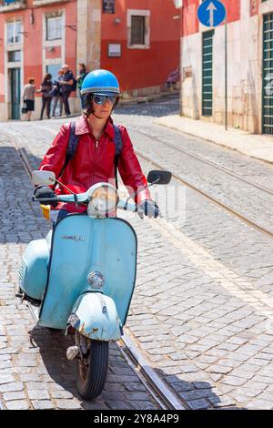 Frau in roter Lederjacke, die auf einem blassblauen Piaggio Vespa Roller in einer Kopfsteinpflasterstraße in Lissabon, Portugal, reitet Stockfoto