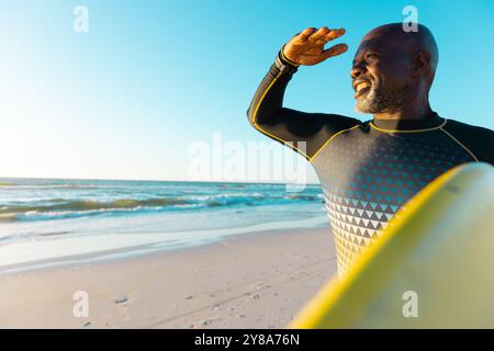 Lächelnder, kahler afroamerikaner, der Augen abschirmt und Surfbrett am Strand gegen den Himmel trägt Stockfoto