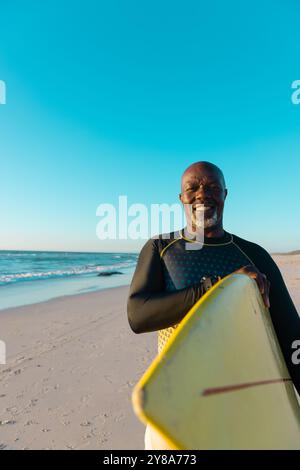 Ein glattes Afroamerikaner lächelt, das Surfbrett am Strand unter blauem Himmel trägt Stockfoto