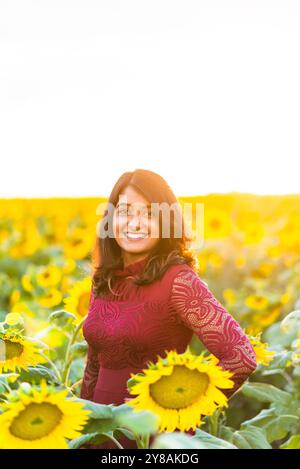Lächelnde Frau, die in einem Sonnenblumenfeld während des Sonnenuntergangs in der goldenen Stunde steht. Stockfoto