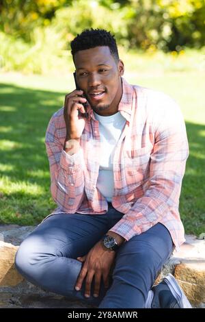afroamerikaner sitzt auf der Treppe und spricht auf dem Smartphone im sonnigen Garten, Kopierraum Stockfoto