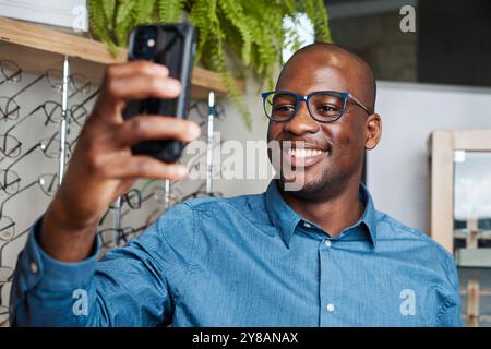Ein lächelnder Optometrist macht ein Selfie im Brillengeschäft mit ausgestellten Brillen Stockfoto