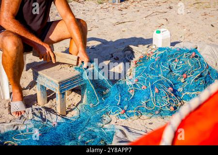 Ein Fischer sitzt auf einem Stuhl am Strand und reinigt Fischernetz von Muscheln, zerschlagen sie mit einem Holzbrett. Stockfoto