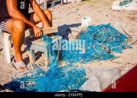 Ein Fischer sitzt auf einem Stuhl am Strand und reinigt Fischernetz von Muscheln, zerschlagen sie mit einem Holzbrett. Stockfoto