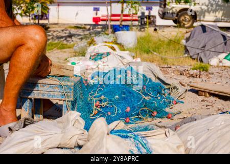 Ein Fischer sitzt auf einem Stuhl am Strand und reinigt Fischernetz von Muscheln, zerschlagen sie mit einem Holzbrett. Stockfoto