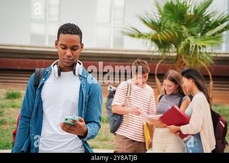Ernsthafter after afroamerikanischer Teenager-Student, der ein Handy auf dem Universitätsgelände benutzt. Ein isolierter Kerl, der schlechte Nachrichten auf seinem Smartphone auf der Highschool sieht. Junger Mann, der draußen mit einem Handy steht. Hochwertige Fotos Stockfoto