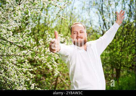 Mann-Allergie genießt nach Behandlung von Saisonallergie im Frühjahr. Porträt eines glücklichen bärtigen Mannes, der im Frühling vor dem Blütenbaum die Daumen nach oben zeigt. Frühjahrsallergiekonzept. Stockfoto