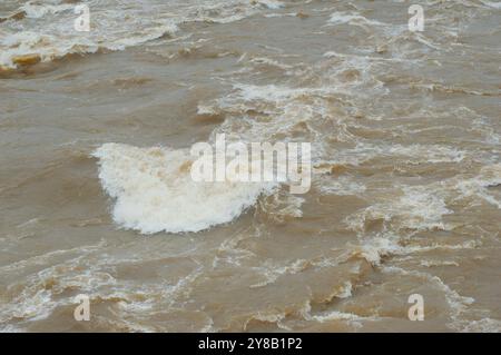 Mittlerer Blick von oben mit Blick auf einen schnellen, sich schnell bewegenden, schlammigen, braunen Fluss, der mit weißen Kappen rollt. Stockfoto