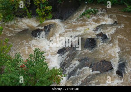 Close Up Overhead Blick hinunter auf die Flussfelsen in einem schnell bewegenden, schlammigen, braunen Fluss, der mit weißen Kappen rollte. Aggresse Stockfoto