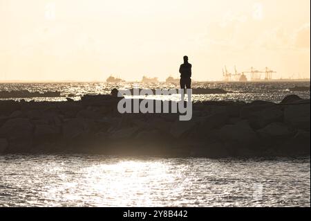 Silhouette eines Mannes, der auf Felsen am Mittelmeer steht und den Sonnenuntergang mit Schiffen am Horizont beobachtet Stockfoto