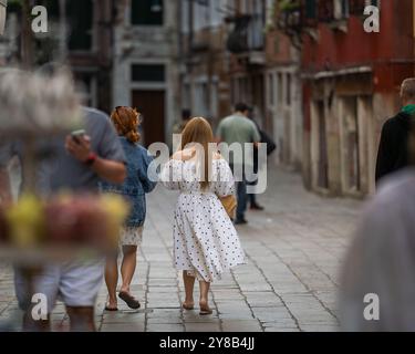 Rückansicht einer Frau in weißem Kleid mit schwarzen Polka Dots in einer Straße in Venedig, Italien. Touristen laufen entlang einer engen Straße in Venedig. Stockfoto