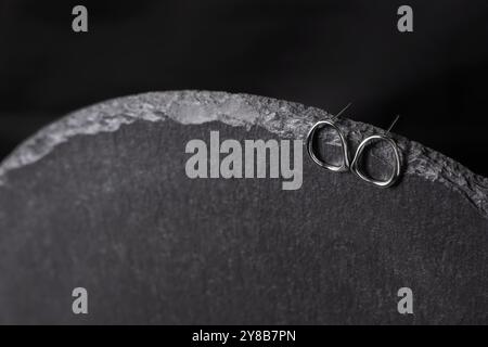 Zwei Ohrringe hängen an einer Keramikschale. Metall-Ohrschmuck auf schwarzem Hintergrund Nahaufnahme. Minimalistischer Schmuck-Ohrring aus Silber. Stockfoto