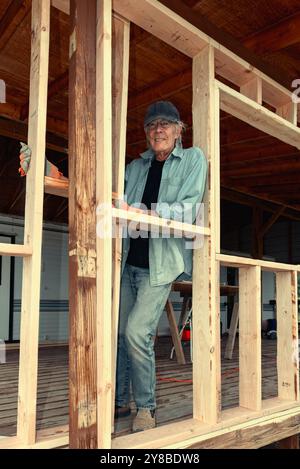 Ein älterer Mann in blau gekleidet steht, lehnt sich an den Rahmen einer Außenwand im Bau, USA. Stockfoto