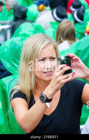 FRÜHE SMARTPHONE, SOMMERSTURM, BEACHVOLLEYBALL, MARIEHAMN, 2011: Eine blonde Frau macht ein Foto mit einem frühen Smartphone. Die Menge vertuscht sich mit Einweg-Regenponchos, während während der Halbfinale im August 2011 bei den PAF Open in Mariehamn, Åland, Finnland, ein riesiger Sommersturm mit einer Regenflut durch das Stadion zieht. Foto: Rob Watkins. INFO: Das PAF Open Beach Volleyballturnier fand zwischen 2009-2013 in Mariehamn, Åland, Finnland statt. Es zog die besten internationalen Teams und Spieler als Rangliste der offiziellen FIVB World Tour an und zeigte hochkarätige Spieler Stockfoto