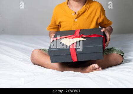 Ein Kind, das auf einem Bett sitzt und eine schwarze Geschenkbox mit einem roten Band hält. Die Box hat ein Etikett mit der Aufschrift: Weihnachten. Das Kind trägt einen Ruf Stockfoto