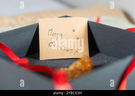 Eine Nahaufnahme einer schwarzen Geschenkbox mit einem roten Band. In der Verpackung befindet sich eine Notiz mit der Aufschrift Frohe Weihnachten!!A in grüner Tinte auf beigefarbenem Papier. Stockfoto