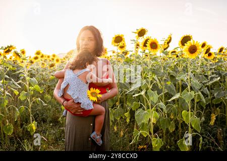 Junge Mutter und kleine Tochter umarmen und spielen zusammen draußen auf einem Sonnenblumenfeld in der Natur. Begriff der Familienqualität Zeit und b Stockfoto