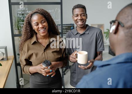 Über die Schulter geschossen von zwei jungen Programmierern, die Tassen Kaffee halten und gemeinsam laut lachen, während sein unerkennbarer Kollege Witze erzählt Stockfoto