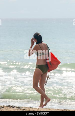 Attraktive, schlanke junge Frau mit grünem Bikini und roter Schultertasche, die an heißen Sommertagen im Mittelmeer entlang eines griechischen Strandes spaziert Stockfoto