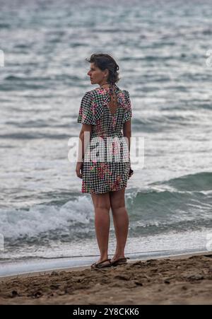 Junge Frau, die allein an einem Sandstrand steht und auf das Meer blickt. Einsame junge Frau, die am Sandstrand am Ufer steht Stockfoto