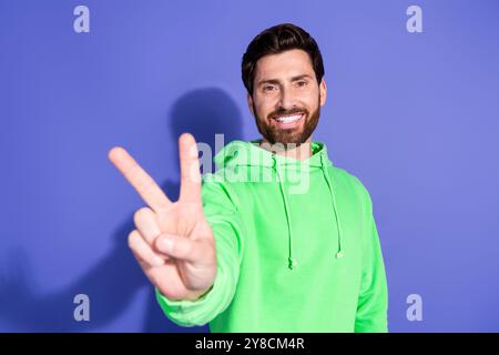 Foto-Porträt des gutaussehenden jungen Mannes zahnig lächelnd, der hellgrünen Hoodie trägt, der in das Kader V-Zeichen zeigt, das auf violettem Hintergrund isoliert ist Stockfoto