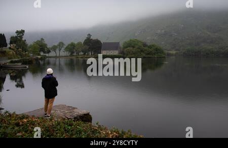 Touristen bewundern die malerische Aussicht auf eine Kirche auf einem See in irland an einem nebeligen Tag. Gougane barra Chuch Stockfoto