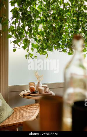 Gemütliche Café-Ecke mit Rattanstuhl, kleinem runden Tisch mit Kaffee, Gebäck und einer Vase mit getrockneten Blumen, umgeben von üppigen hängenden Pflanzen Stockfoto